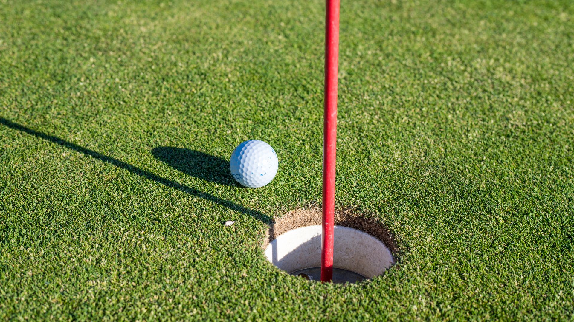 A golf ball rests near the edge of a hole on a putting green, with a red flagstick casting a shadow across the grass—capturing one moment in how long does golfing take during a round.